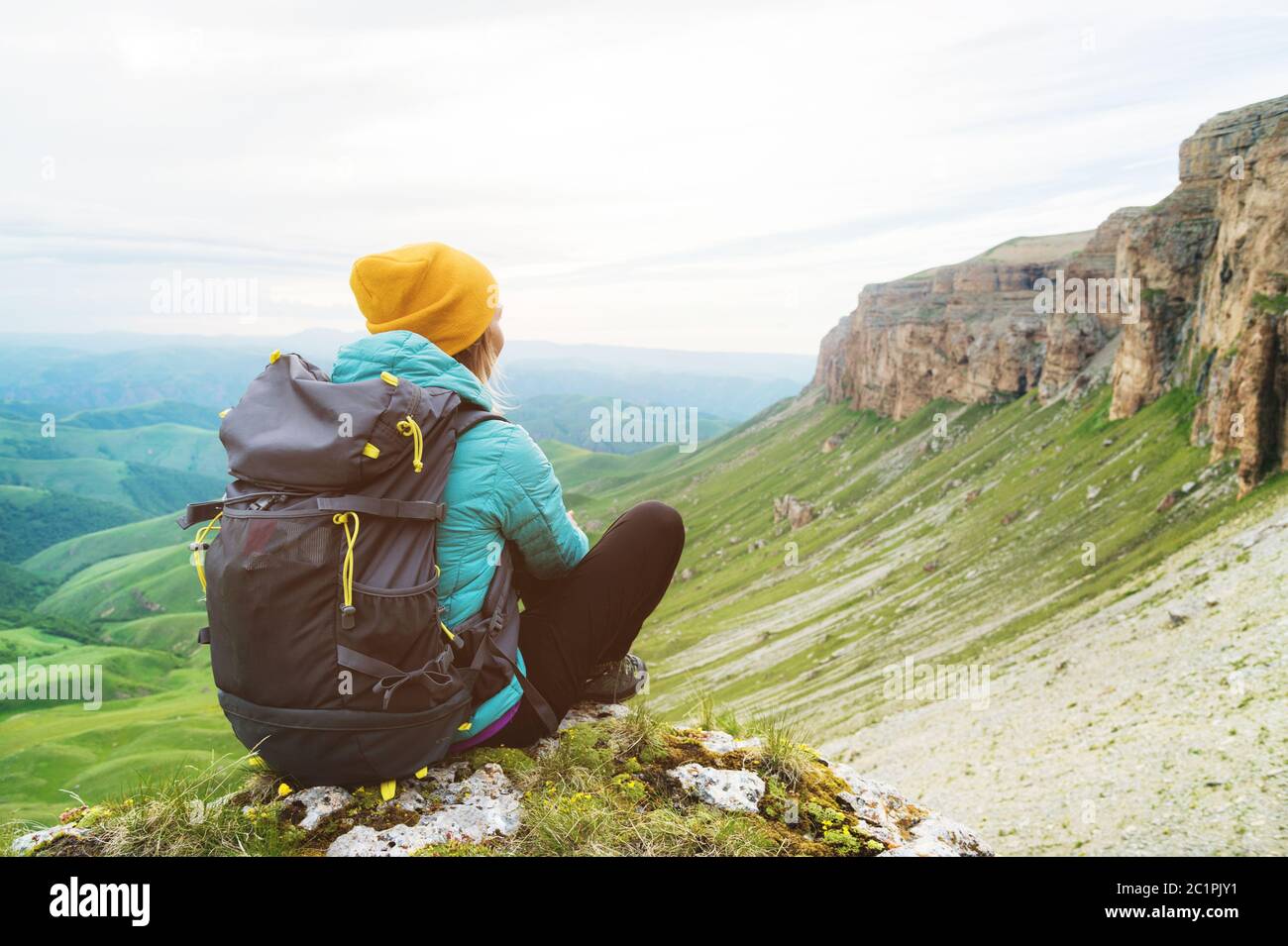 Sitzende zurück Mädchen Reisenden in einem gelben Hut und eine Sonnenbrille sitzen am Fuße der epischen Felsen mit einem Rucksack nächsten und Stockfoto