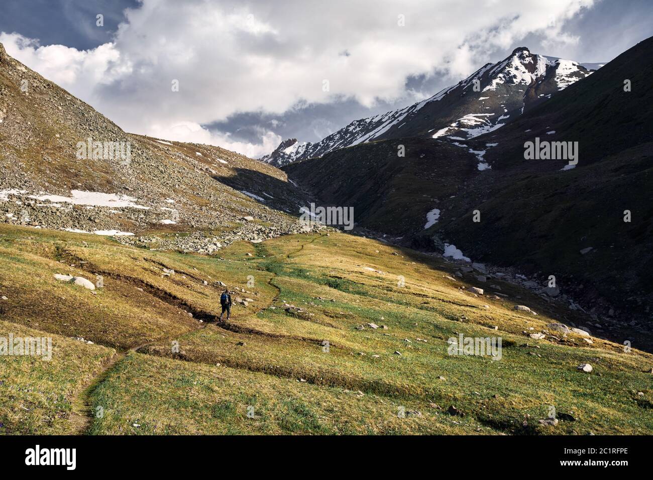 Kleiner Tourist mit Rucksack ist Wandern in der grünen Bergtal mit schneebedeckten Gipfeln und bewölktem Himmel Hintergrund Stockfoto