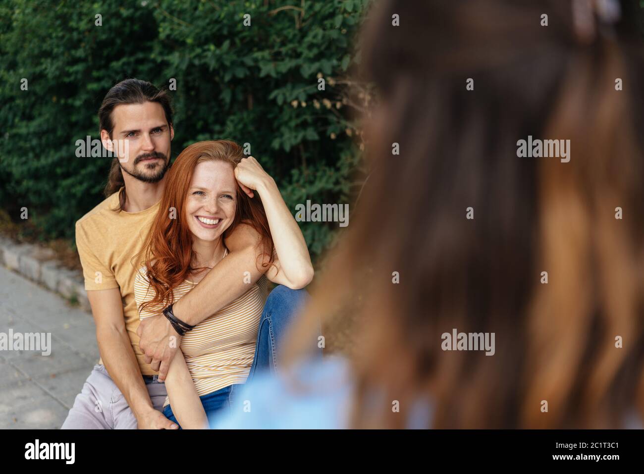 Liebevolles junges Paar in einer engen Umarmung in einem grünen Garten im Gespräch mit einem Freund in einer über die Schulter-Ansicht Stockfoto