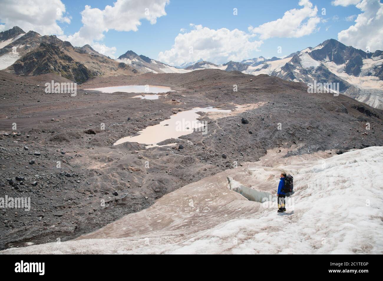 Ein Bergsteiger mit Rucksack geht in Steigeisen entlang eines staubigen Gletschers mit Gehwegen in den Händen zwischen Rissen in der Stockfoto