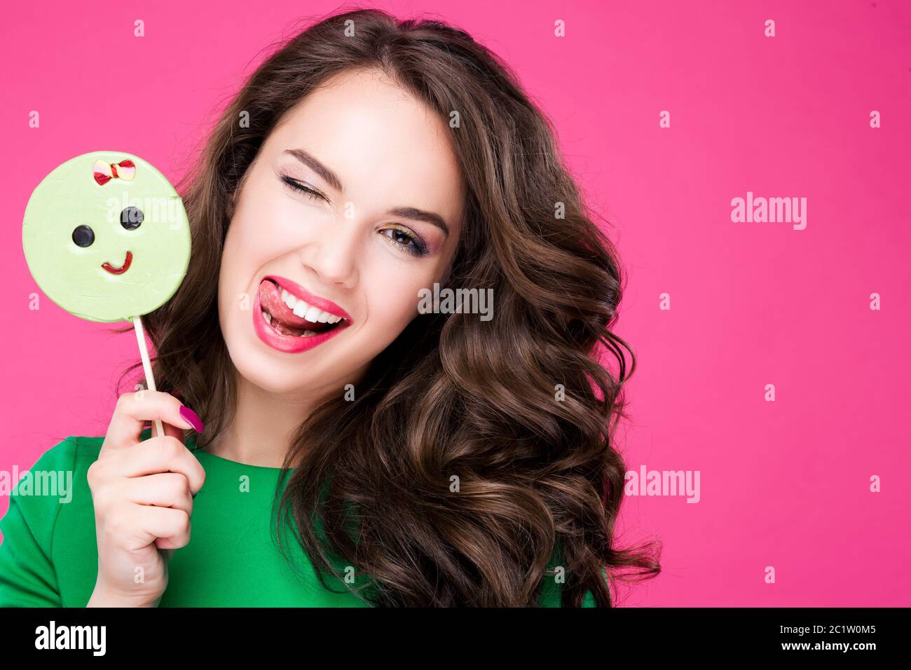 Fröhliche hübsche Frau mit einem Candy zwinkert lacht zeigt Sprache. Brunette Mädchen modisch und stilvoll auf rosa Hintergrund in Stockfoto