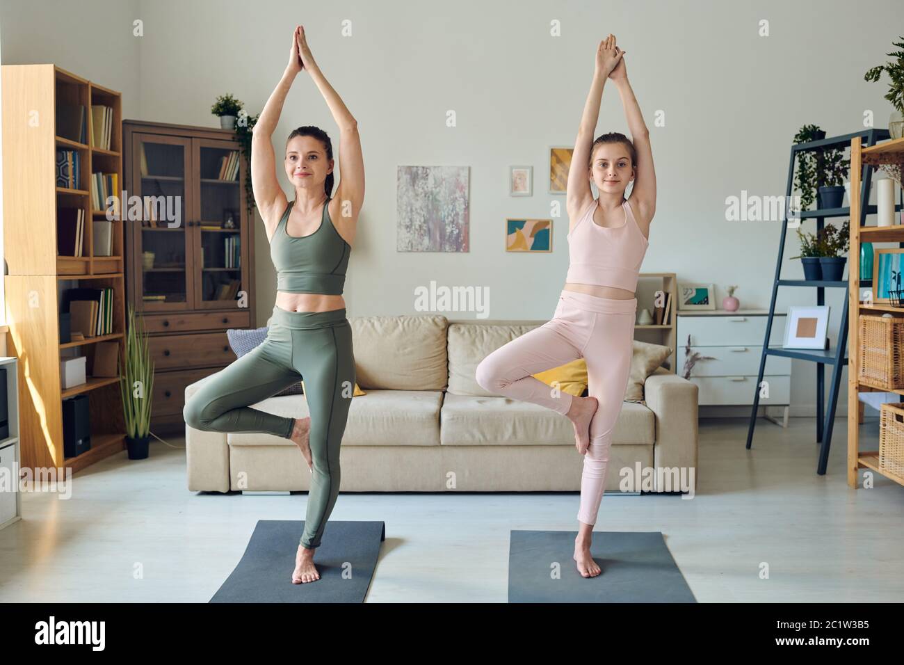 Energische Mutter und ihre Teenager-Tochter balancieren auf einem Bein, während sie im Wohnzimmer Baumpose tun Stockfoto