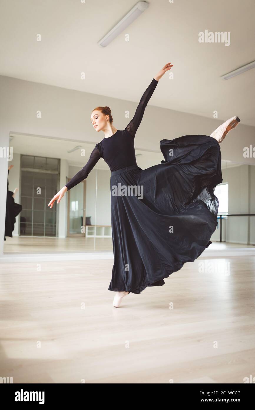 Kaukasische Balletttänzerin in einem schwarzen Kleid und konzentriert sich auf ihre Übung in einem hellen Studio Stockfoto