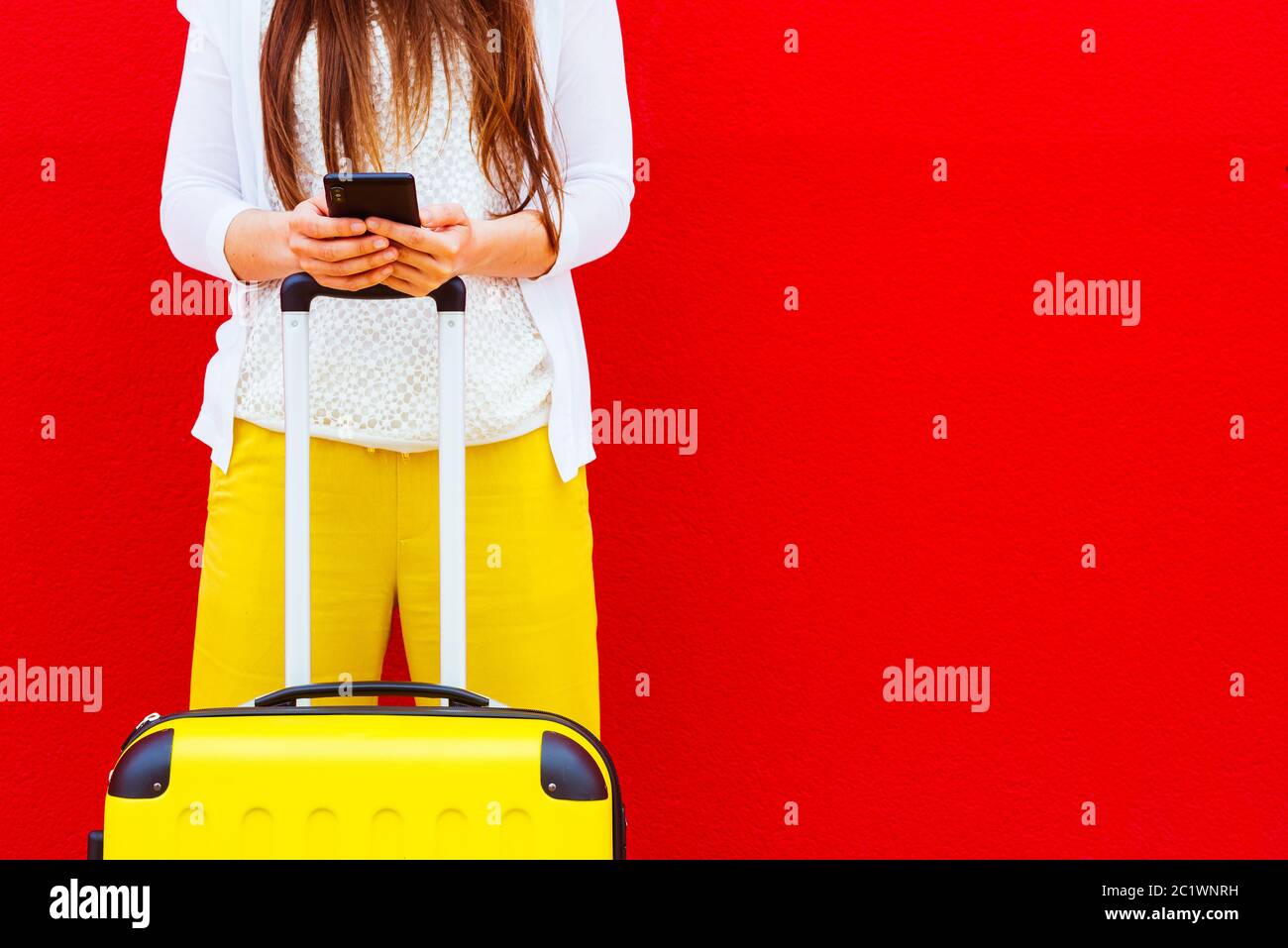 Frau mit einem gelben Koffer, die Nachrichten auf ihrem Handy liest, das sich an eine rote Wand lehnt Stockfoto