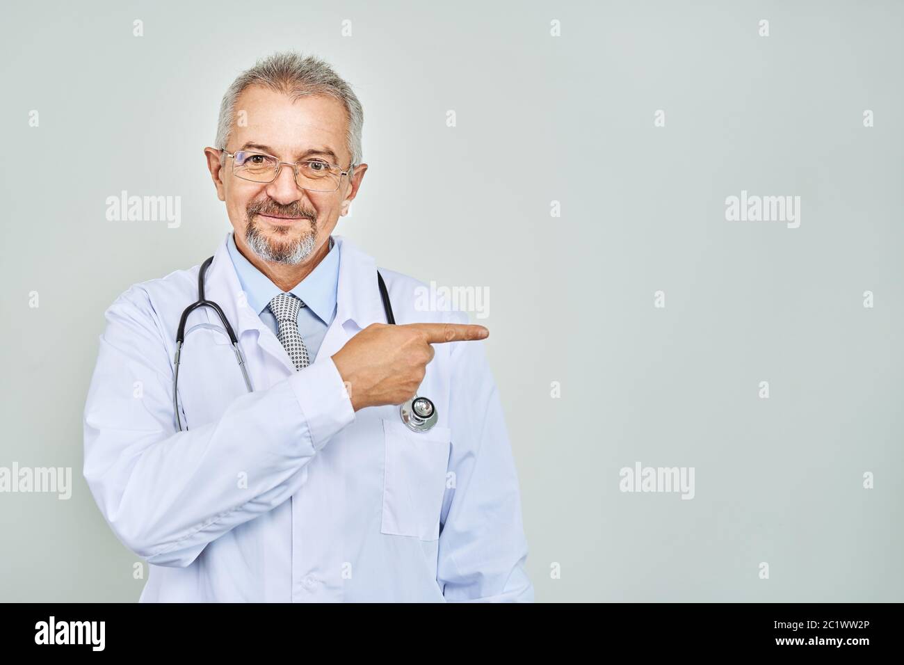 Fröhliche reife Arzt posing und lächelnd an Kamera, Gesundheitswesen und Medizin. Auf blauem Hintergrund isolieren. Hand bei copy Raum zeigt. Stockfoto