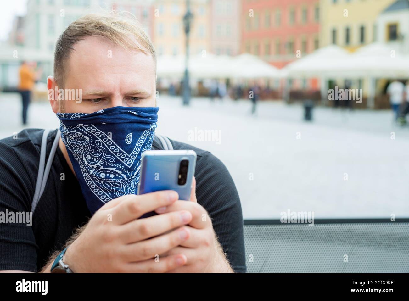 Erwachsene Mann in Bandana als Gesichtsmaske nutzt Smartphone in Stadt Café Terrasse. Solo Outdoor-Aktivitäten. Sicherheit Stadtspaziergang Stockfoto