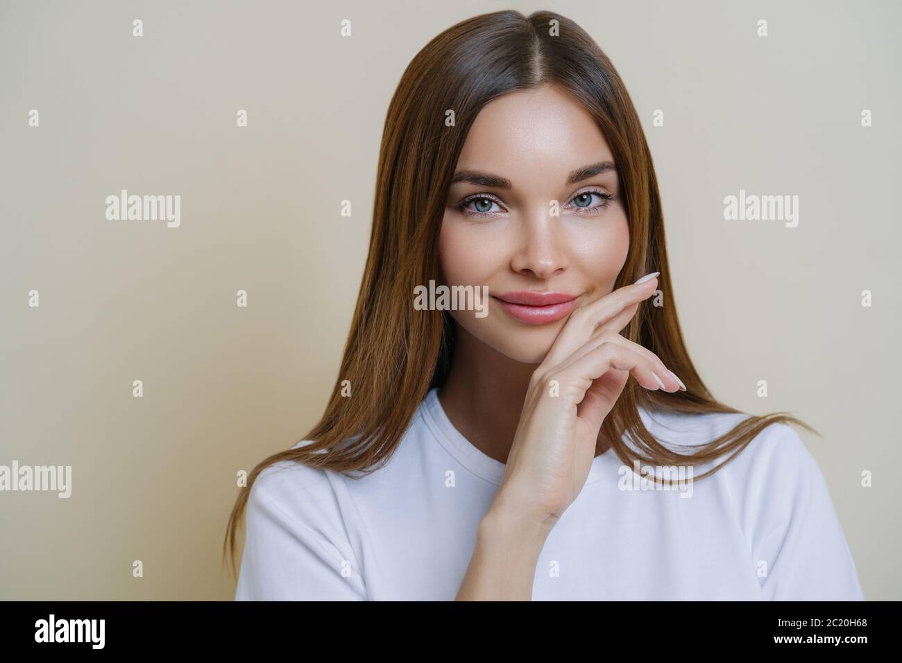 Nahaufnahme der charmanten dunkelhaarigen Frau hält Hand auf Wange, in lässigen weißen T-Shirt gekleidet, hat minimale Make-up, stellt innen isoliert über beige Stockfoto