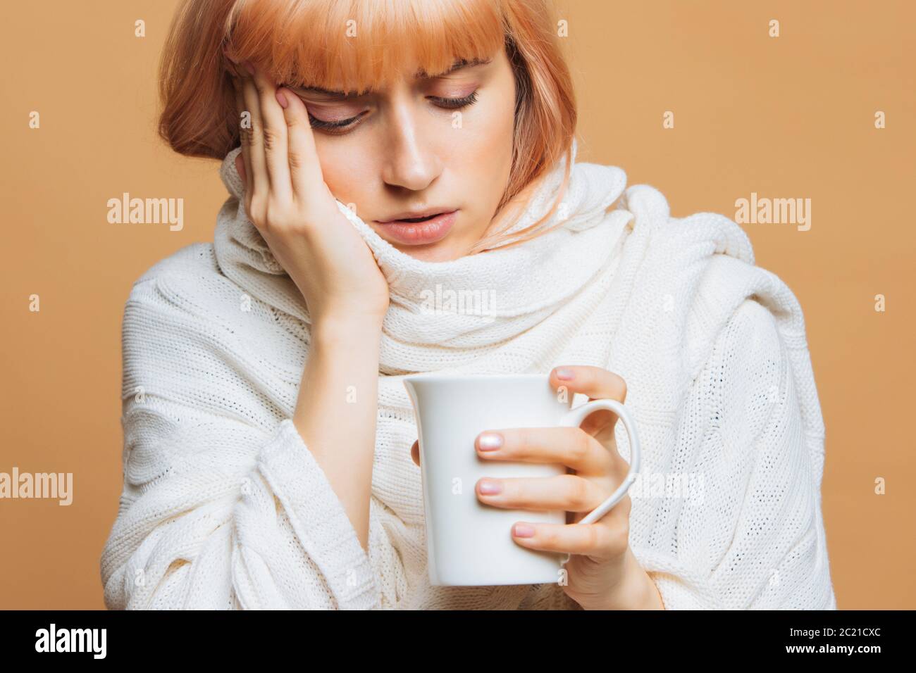 Sickness nette junge Frau mit Erdbeer blonden Haaren, warm karierten mit Kopfschmerzen, hält Tasse heißen Tee, berühren ihre Stirn, Gefühl der ersten symp Stockfoto