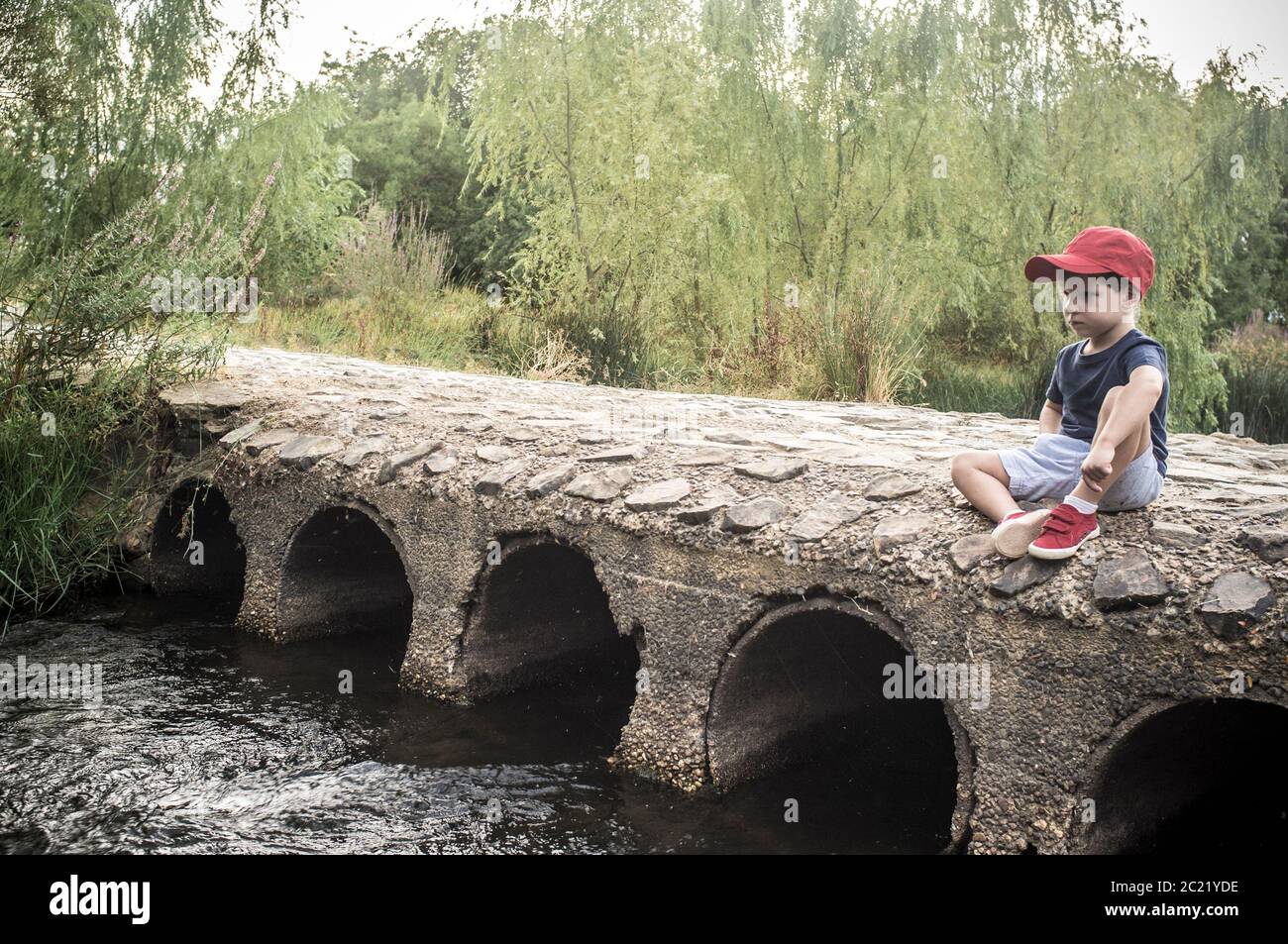 Kleiner Junge sitzt über Brücke und beobachtet, wie das Wasser fließt. Natur für Kinder entdecken Konzept Stockfoto