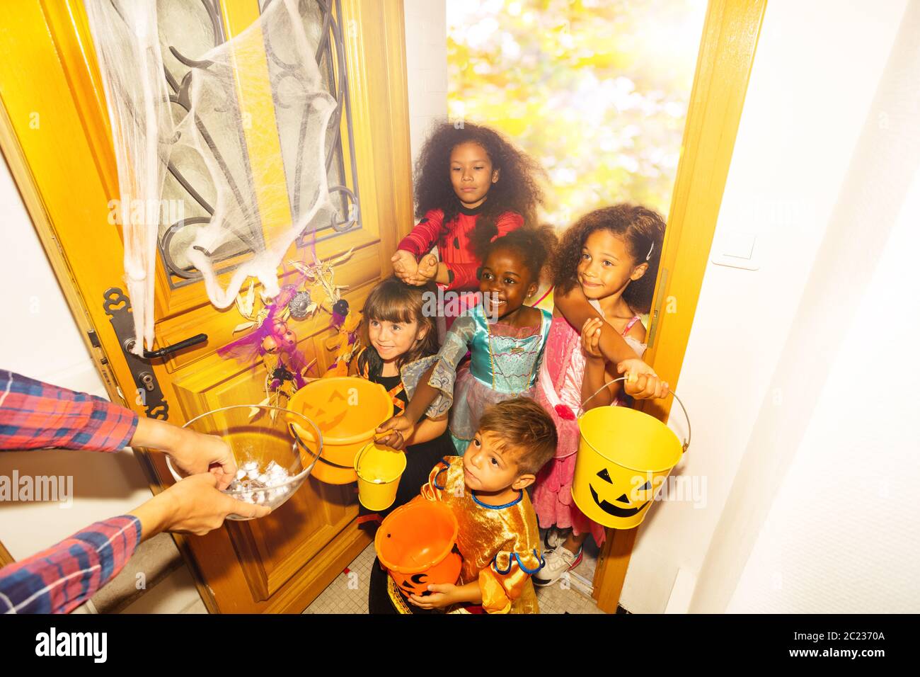 Viele Kinder vielfältig aussehende Jungen und Mädchen in Halloween-Kostümen stehen in der Tür mit orangefarbenen Eimern für Süßigkeiten erhalten Süßigkeiten von Erwachsenen Stockfoto