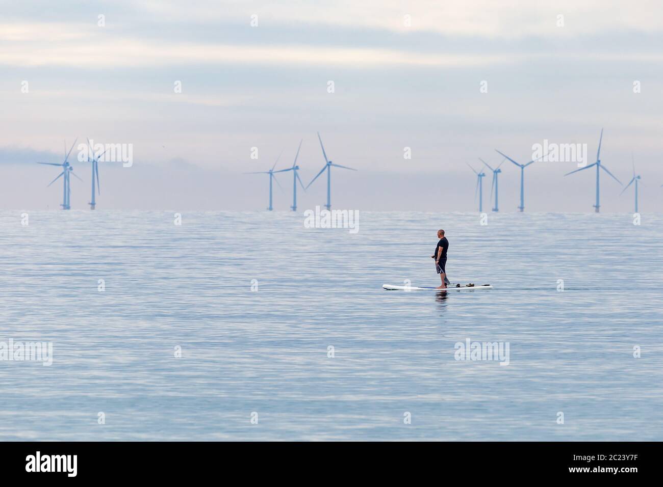 Worthing, Sussex, UK; 15. Juni 2020; Mann auf dem Paddleboard Gleiten durch ein ruhiges Meer. Es gibt weiches Morgenlicht. Eine Reihe von Windenergieanlagen sind Stockfoto