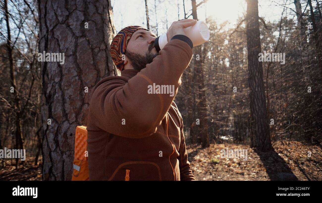 Ein junger müde Kaukasusmann trägt eine Pause mit einem Rucksack sitzen und Trinkwasser auf einem Baum in einem Naturwald während Stockfoto