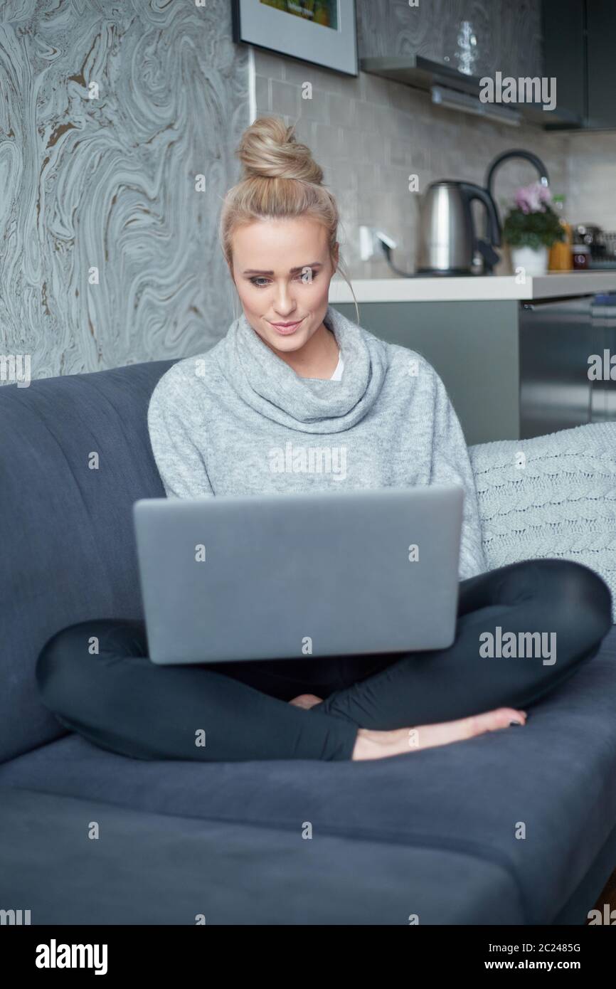 Fröhliche junge blonde Frau sitzt auf der Couch im Wohnzimmer und mit Laptop Stockfoto