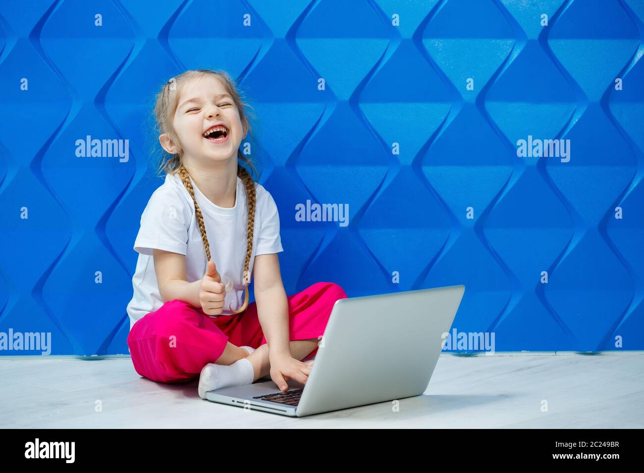 7 Jahre altes Mädchen in einem weißen T-Shirt sitzt mit einem Laptop auf dem Boden und drückt die Tasten Stockfoto