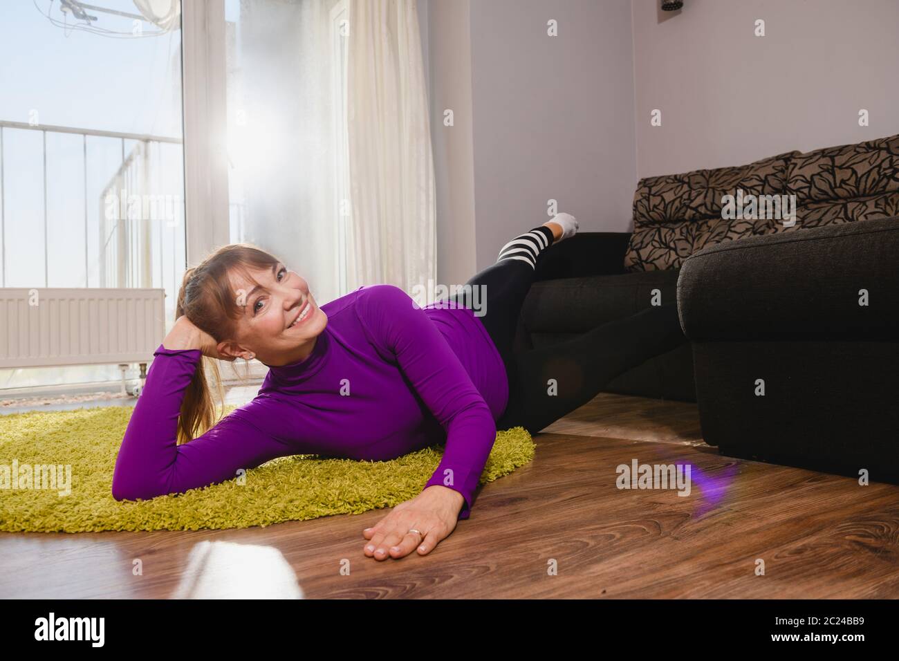 Lächelnd nette alte Frau hat Fitness-Training drinnen, heben die Beine auf der Couch und lag auf dem Boden Teppich in Sport Leggings und lila Rollkragen, Senior Aktivität Stockfoto