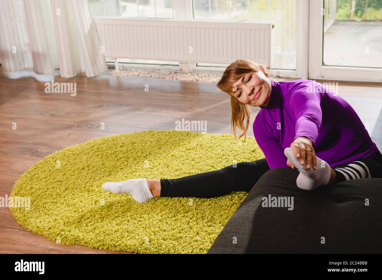 Ältere Frau Rentner machen Workout Stretching Übung zu Hause Sofa und Teppich, tragen Sport Leggings und lila Top, aktiven Ruhestand Lebensstil Stockfoto