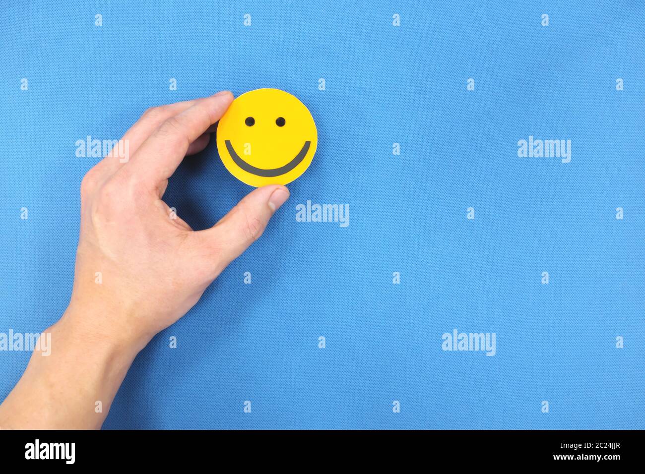 Glück und Positivität Konzept. Hand hält gelb lächelndes Gesicht in blauem Hintergrund mit Kopierraum. Stockfoto