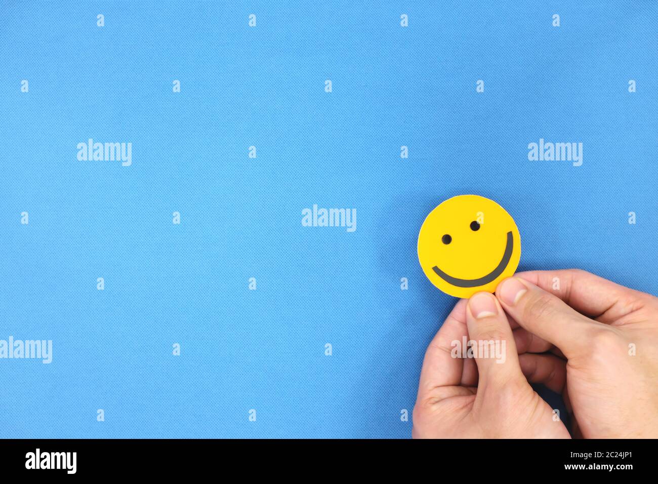 Glück und Positivität Konzept. Hand hält gelb lächelndes Gesicht in blauem Hintergrund mit Kopierraum. Stockfoto