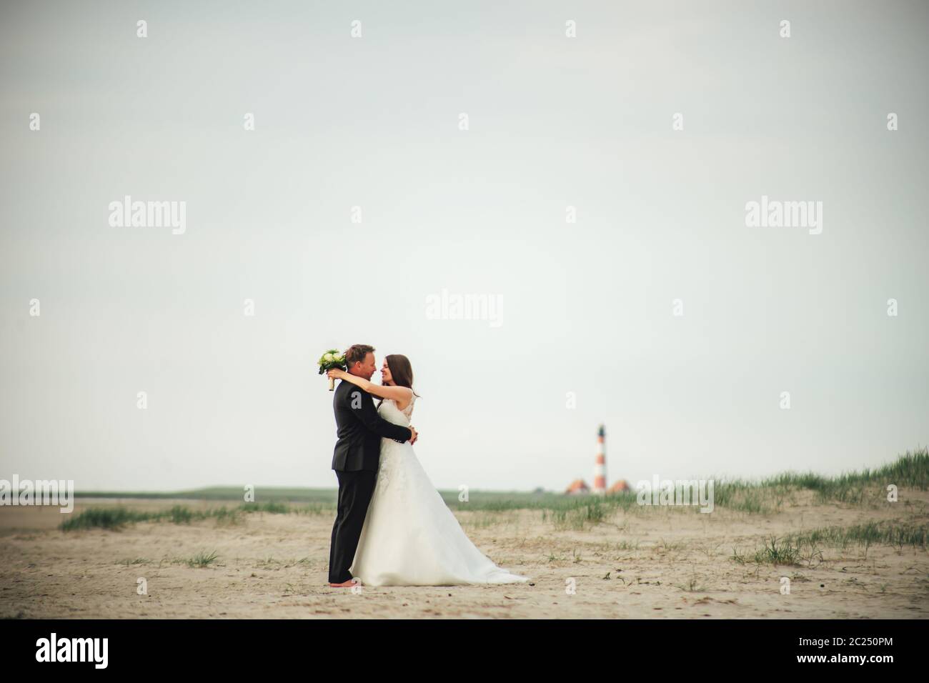 Brautpaar steht und umarmt am Sandstrand. Braut und Bräutigam schauen sich gegenseitig an und lächeln. Verschwommener Leuchtturm im Hintergrund. Hochzeitstag Stockfoto