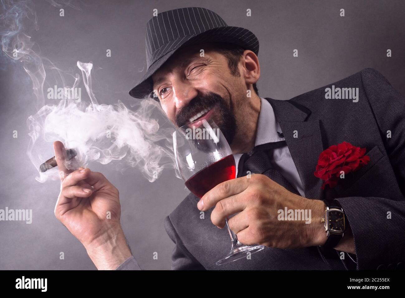 Der lächelnde zwinkende elegante Gentleman in Anzug und Hut raucht Zigarre und trinkt Wein auf grauem Hintergrund. Stockfoto