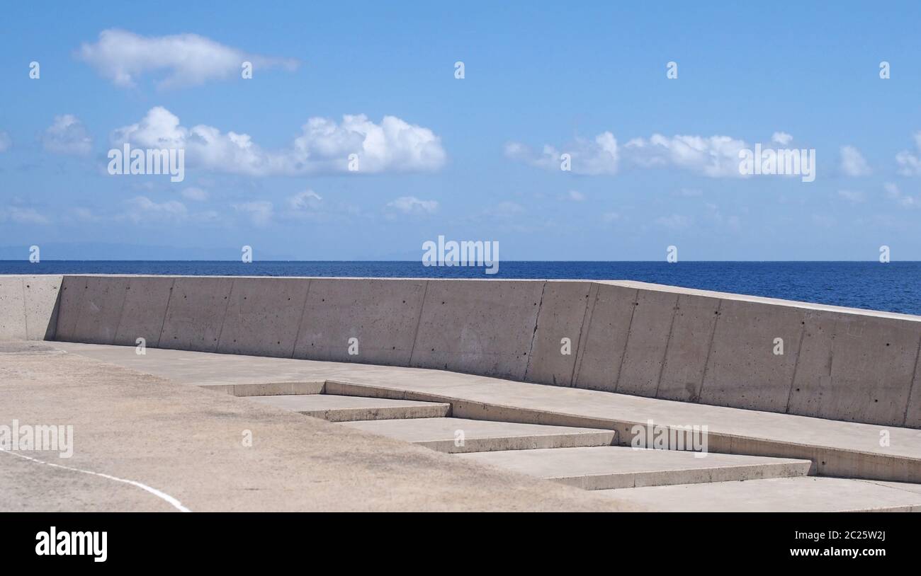 Langes Bild einer Betonwand mit Stufen gegen ein ruhiges blaues Meer und sonnenbeschienenen blauen Himmel Stockfoto