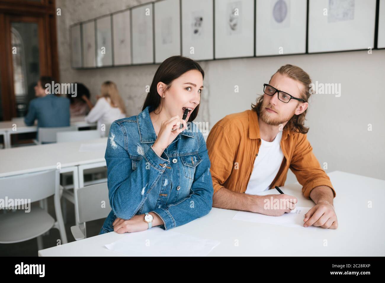 Hübsches Mädchen beißen Stift während im Klassenzimmer sitzen und versuchen, schreiben Sie aus Test in Junge, der es bemerkt. Junger Mann sitzt am Schreibtisch im Klassenzimmer und Stockfoto