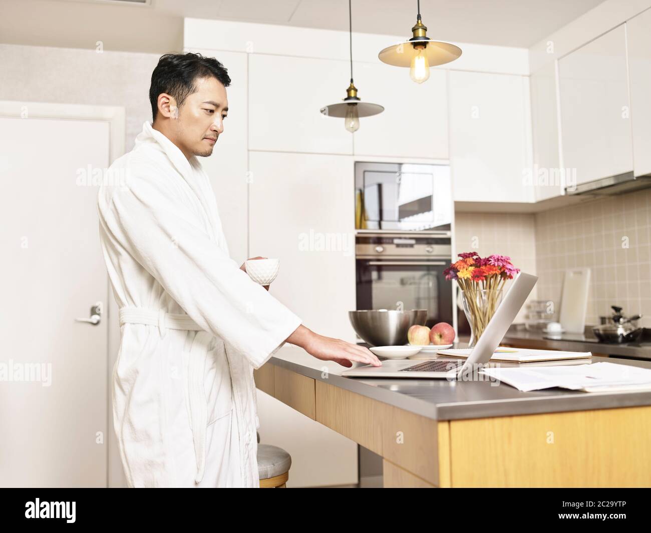 Junger asiatischer Geschäftsmann im Bademantel, der von zu Hause aus an der Küchentisch steht und mit einem Laptop eine Tasse Kaffee hält Stockfoto