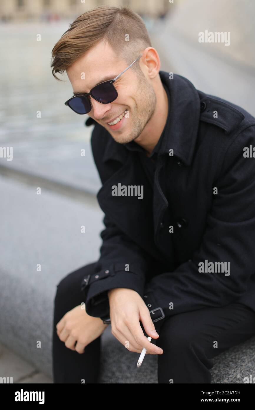 Junger lächelnder Mann in schwarzer Jacke sitzt auf Louvre Pyramide und rauchen. Stockfoto