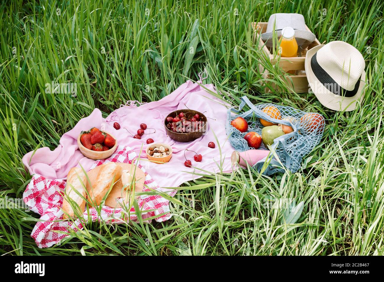 Zero Waste Sommer Picknick auf dem mit Kirschen in den hölzernen Kokosnussschalen, frischem Brot und Glasflasche Saft oder Smoothie auf rosa Decke, Flatlay Stockfoto