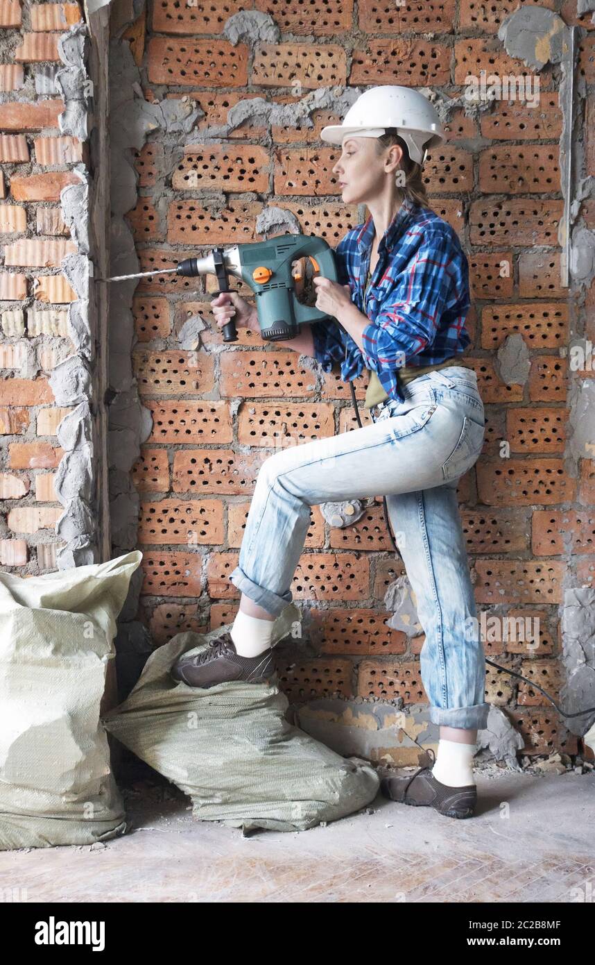 Junge Frau in Arbeitskleidung und ein Schutzhelm steht neben einer Ziegelwand und macht ein Loch Witz Stockfoto