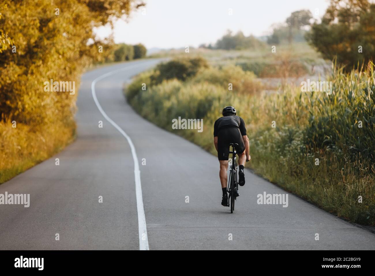 Rückansicht eines starken Radfahrers mit sportlicher Körperform auf der asphaltierten Straße zwischen Bäumen und grünen Büschen in schwarzem Schutzhelm und sp Stockfoto