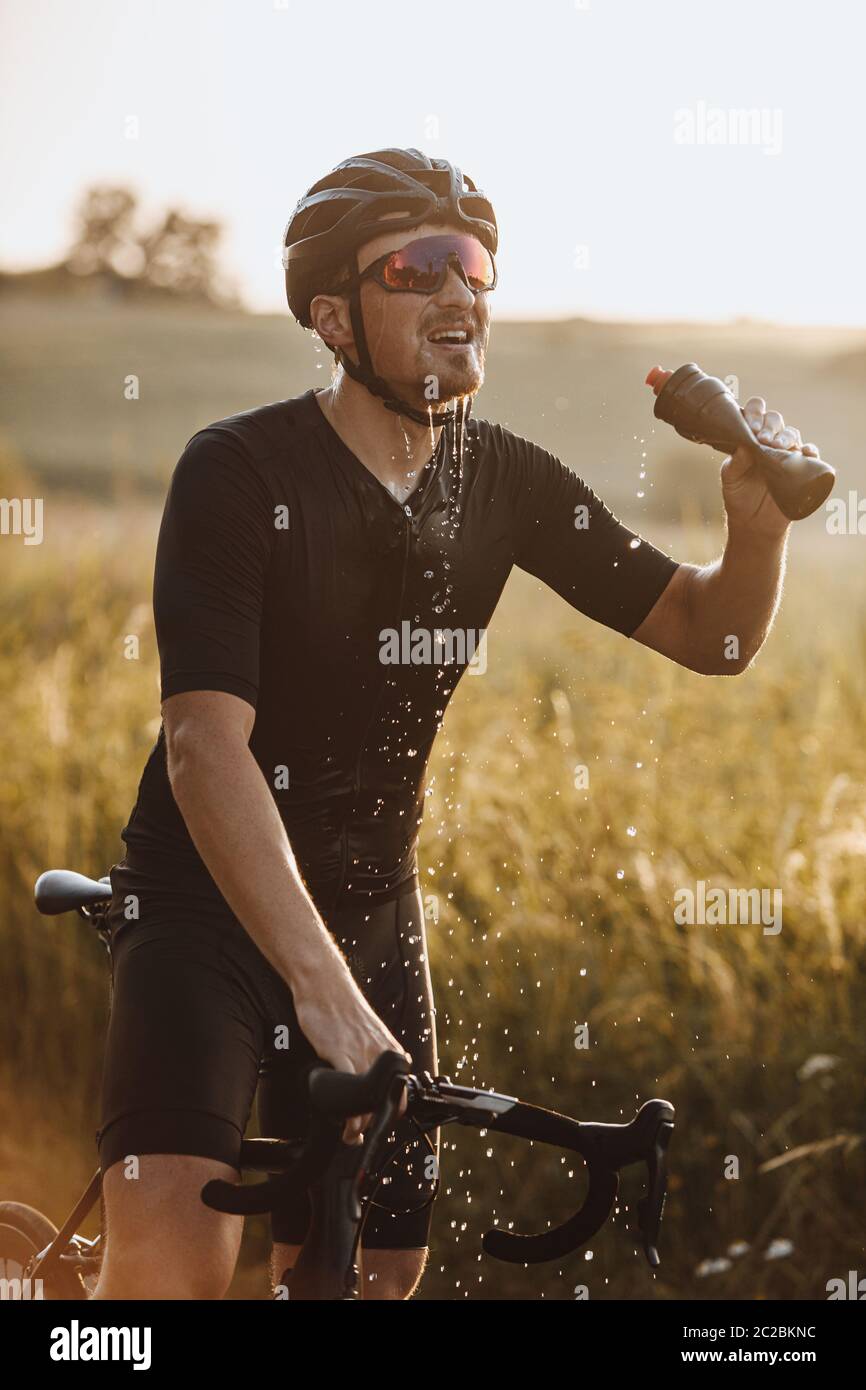 Starker bärtiger Mann in aktiver Kleidung, Schutzhelm und Sportbrille spritzt frisches Wasser aus der Flasche im Freien. Müde Radfahrer nach Durst Stockfoto