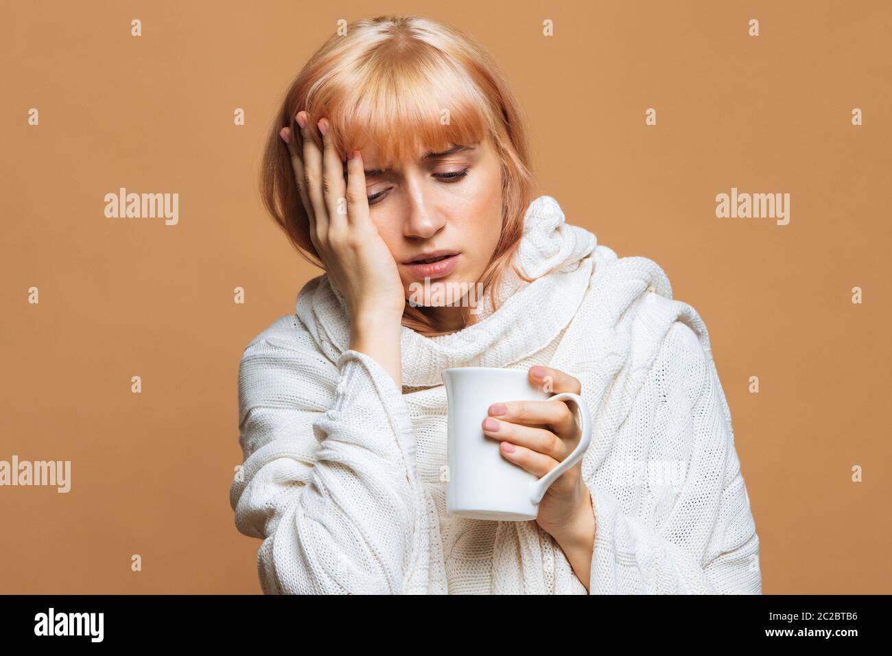 Sickness nette junge Frau mit Erdbeer blonden Haaren, warm karierten mit Kopfschmerzen, hält Tasse heißen Tee, berühren ihre Stirn, Gefühl der ersten symp Stockfoto