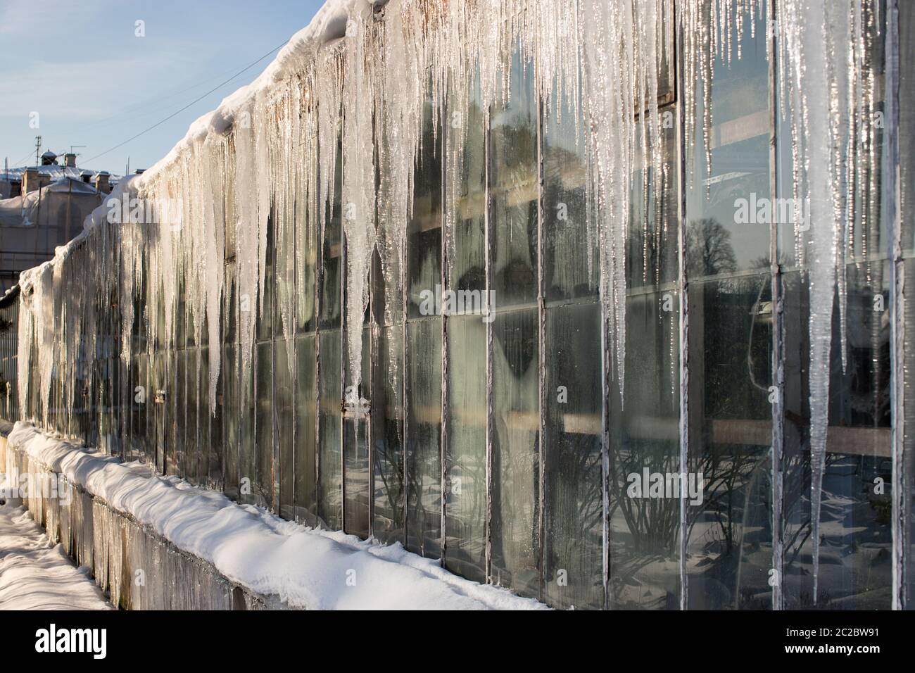Scharfe Eiszapfen auf dem Dach des Gewächshauses in sonnigen Wintertag auf der Sonnenseite, horizontal. Bildung von Eiszapfen im Tauwetter. Eiszapfen hängen nach unten Fr. Stockfoto