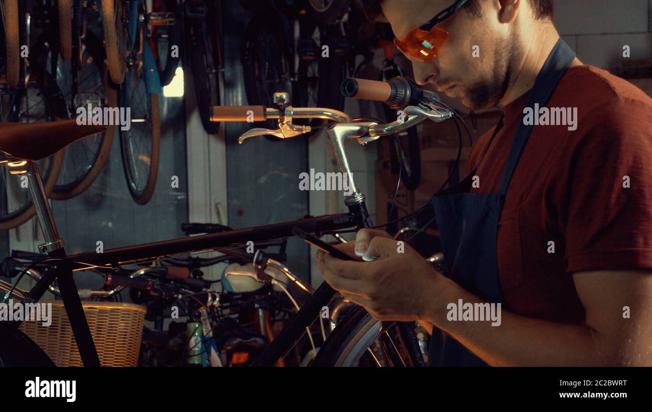 Thema kleine Unternehmen Fahrrad Reparatur. Junge kaukasische Brünette Mann trägt Schutzbrille, Handschuhe und fartukhe nutzt Handy tec Stockfoto