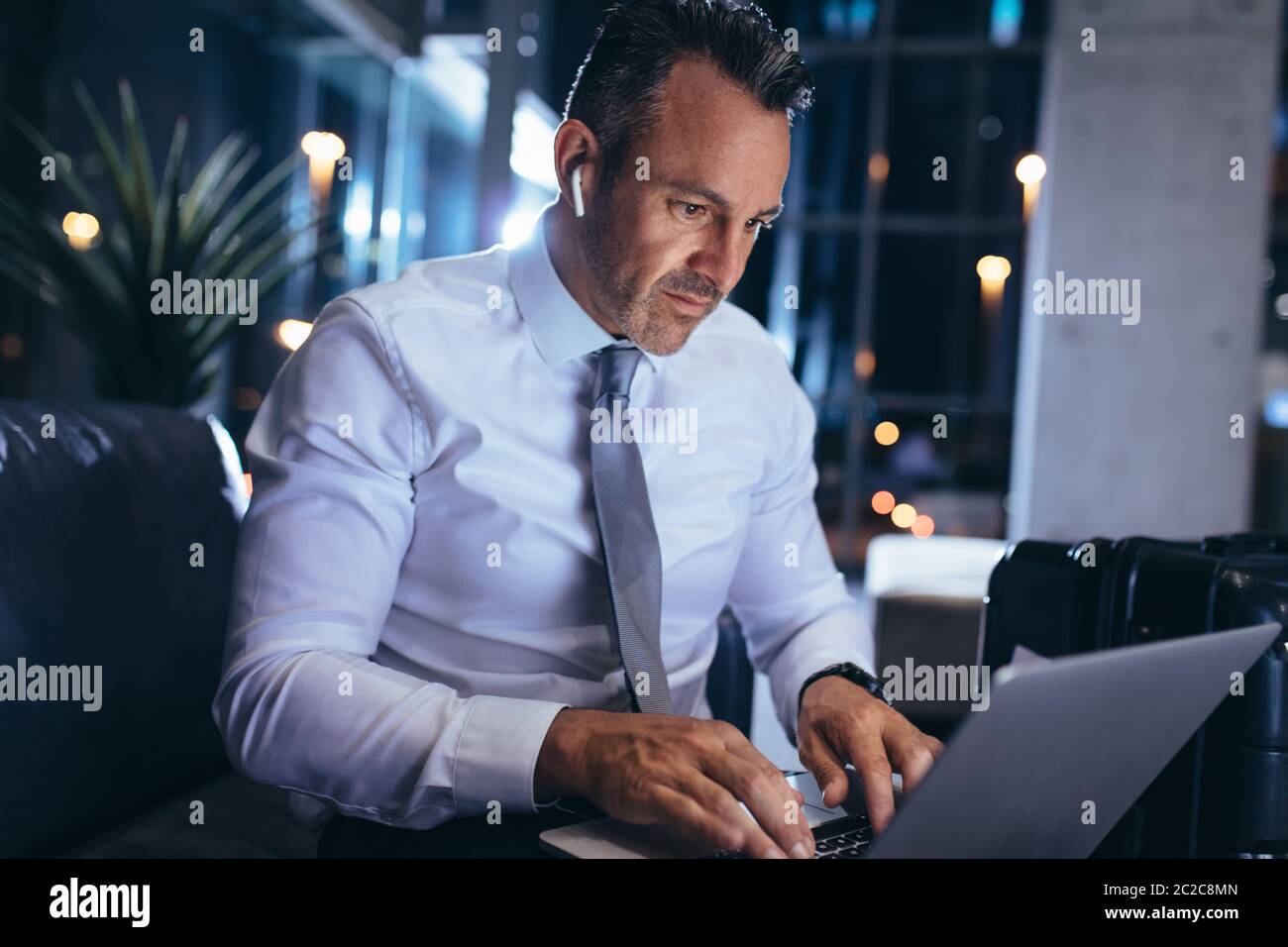 Ein reifer Geschäftsmann mit kabellosen Kopfhörern, die in der Flughafenlounge mit einem Laptop sitzen. Geschäftsprofi liest E-Mails auf dem Laptop, während Sie Stockfoto