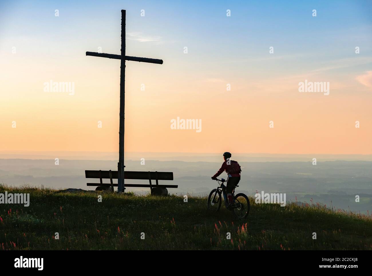 Hübsche ältere Frau, die mit ihrem Elektro-Mountainbike in warmen Morgensonnen unter dem Gipfelkreuz von Salmas hoch über Oberstaufen, Allgauer Alpen, Binnen fährt Stockfoto