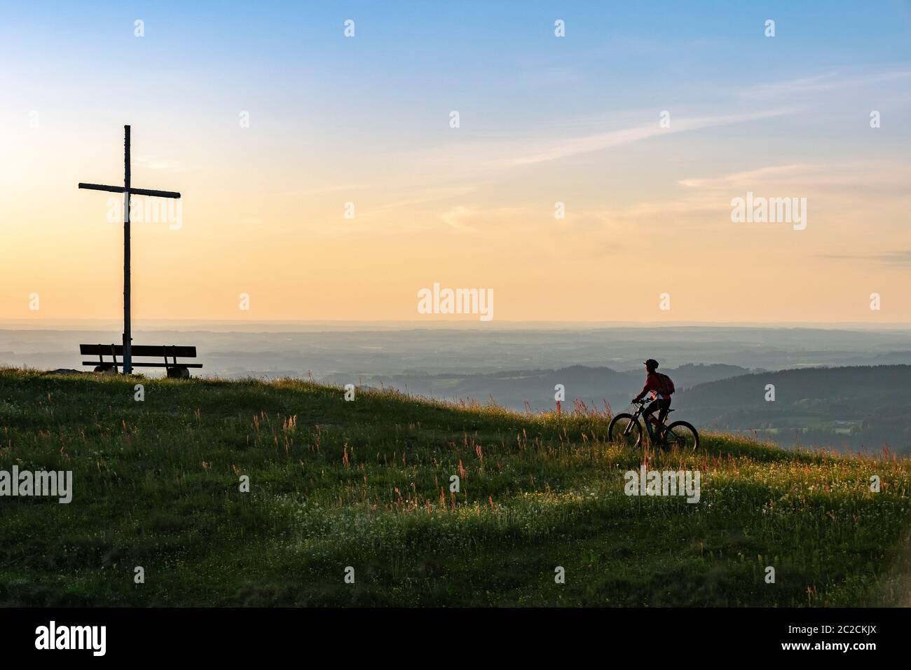 Hübsche ältere Frau, die mit ihrem Elektro-Mountainbike in warmen Morgensonnen unter dem Gipfelkreuz von Salmas hoch über Oberstaufen, Allgauer Alpen, Binnen fährt Stockfoto