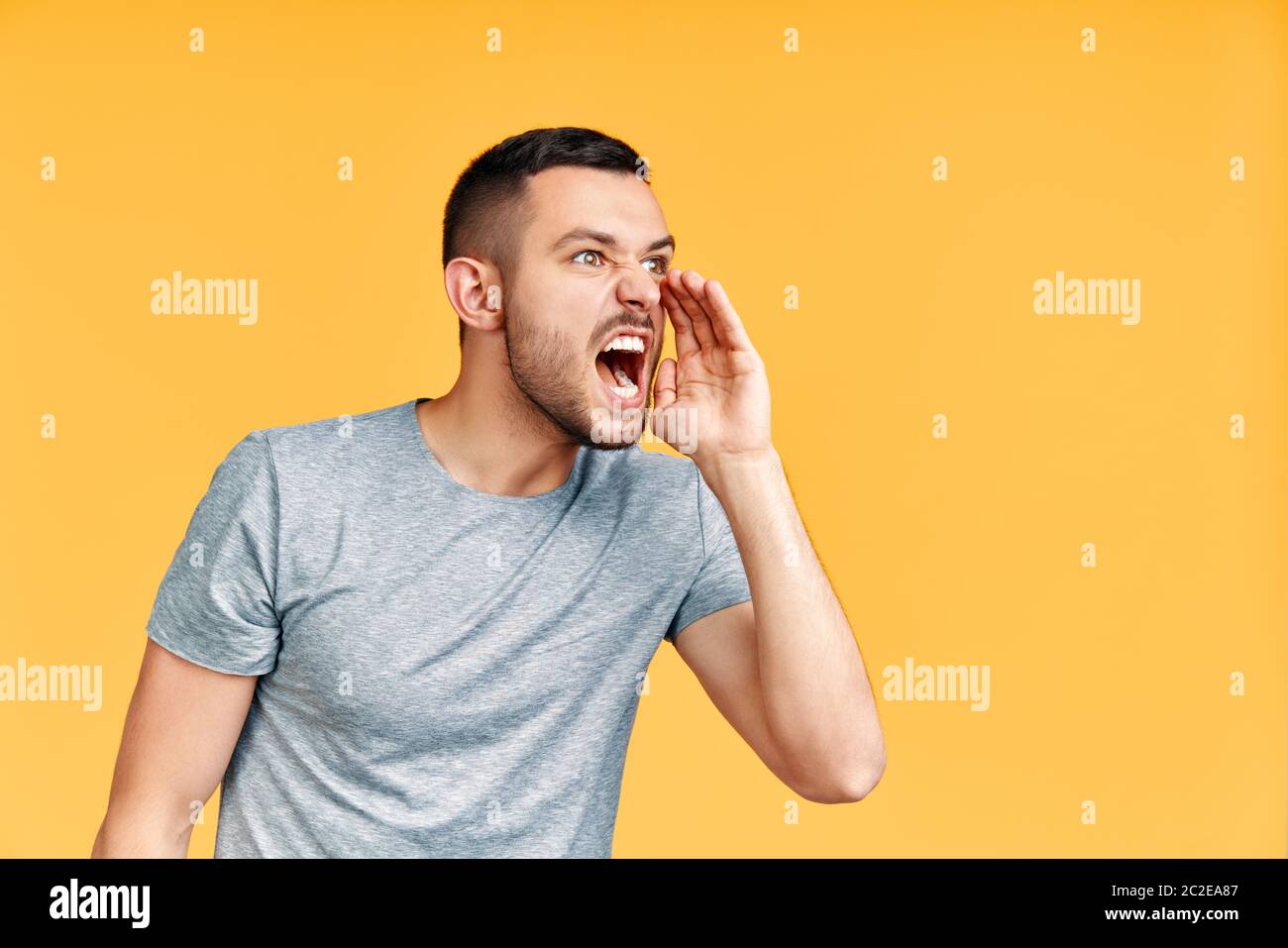 Junger wütender Mann, der laut und laut auf Seite mit der Hand auf dem Mund schreit und Platz für Text auf gelbem Hintergrund kopiert Stockfoto