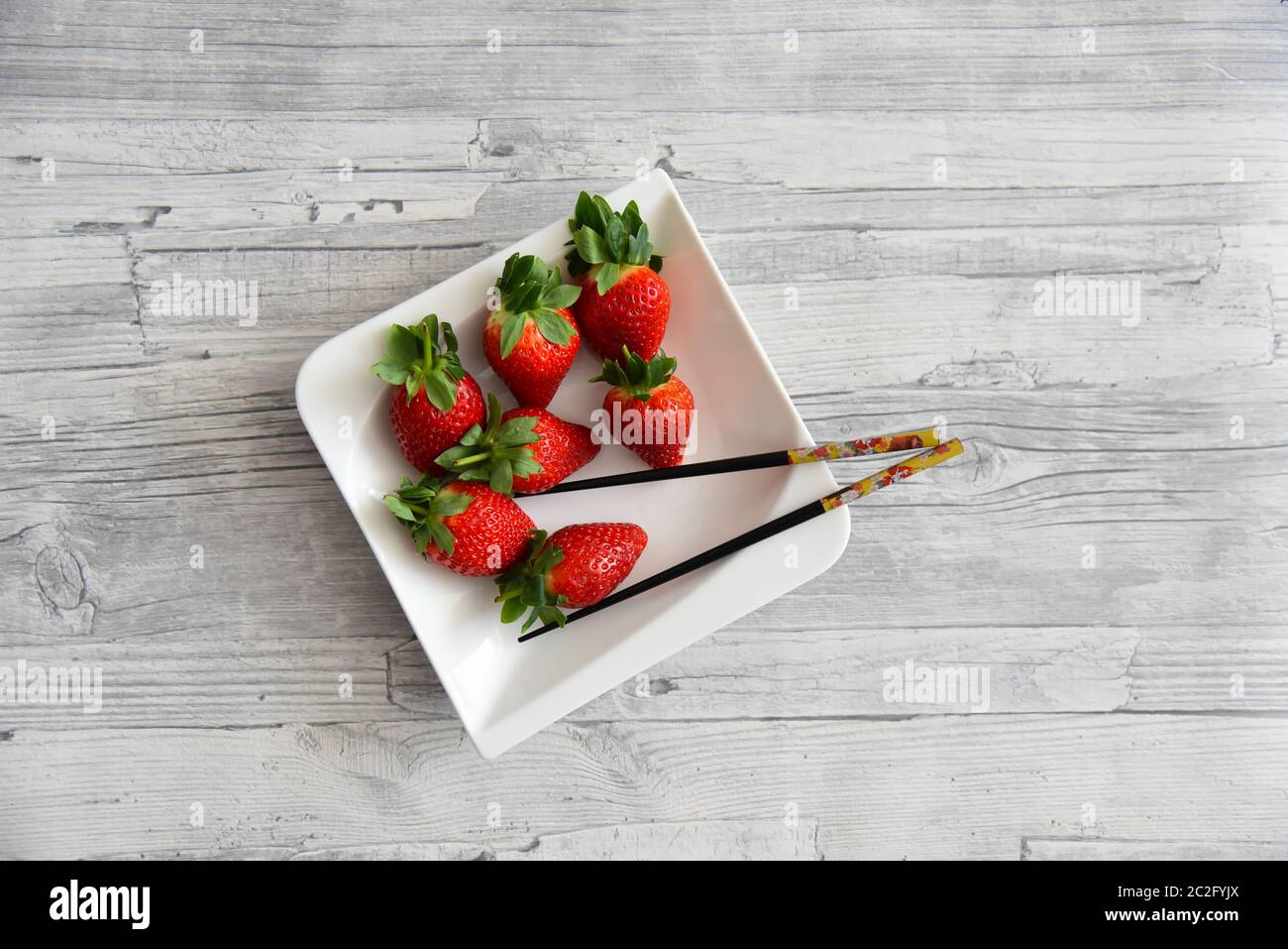 Saftige rote Erdbeeren mit grünen Blättern auf einem weißen Teller mit Essstäbchen. Holztisch Hintergrund. Stockfoto