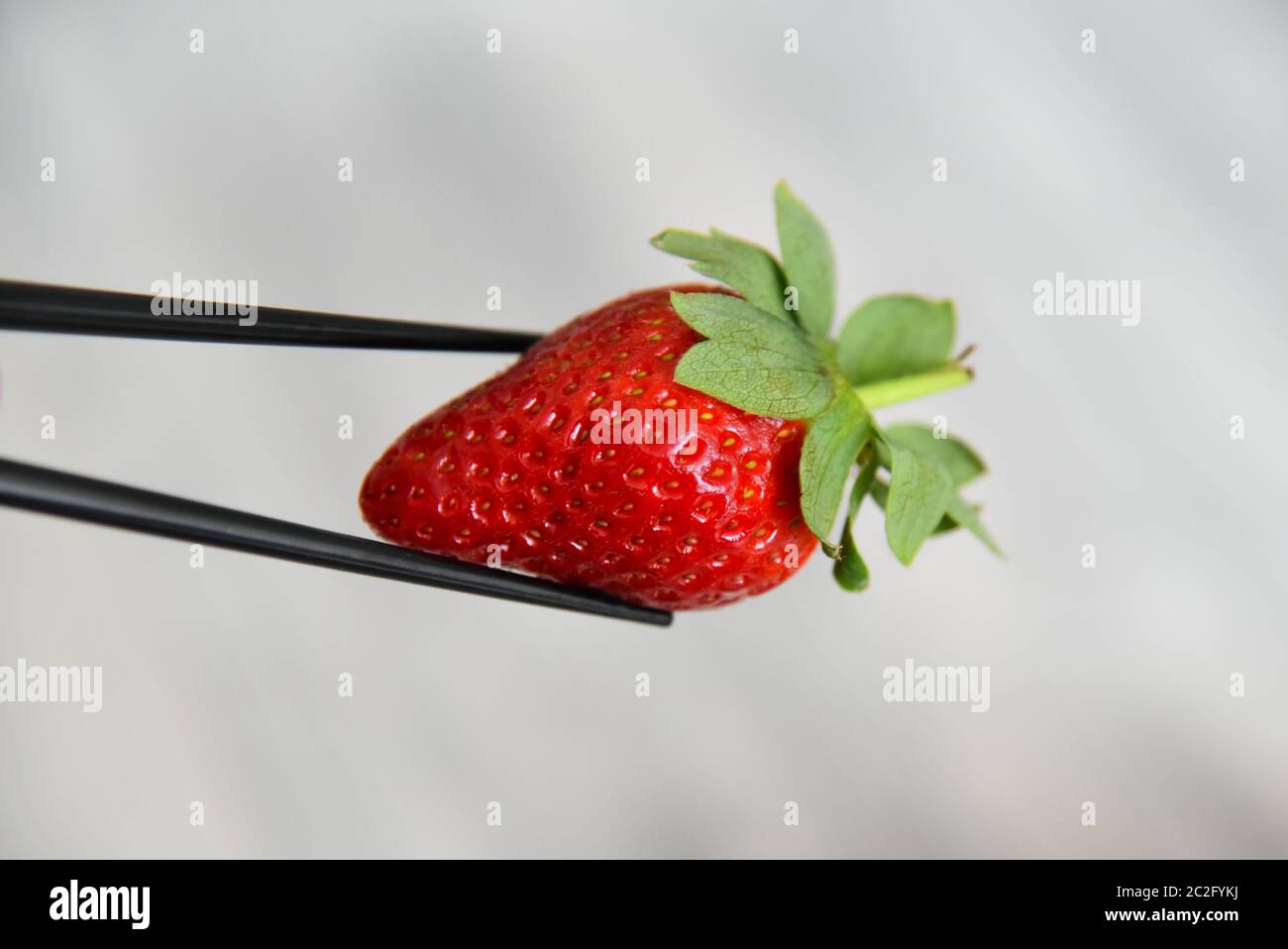 Nahaufnahme von Essstäbchen mit einer roten, saftigen Erdbeere mit grünen Blättern vor einem verschwommenen Hintergrund. Stockfoto