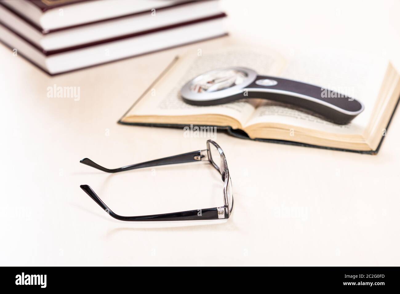 Lesen Buch mit Low Vision - Brille und Lupe auf offenen Buch in der Nähe von Stapel von Büchern über blasse Tabelle (Schwerpunkt im Vordergrund) Stockfoto