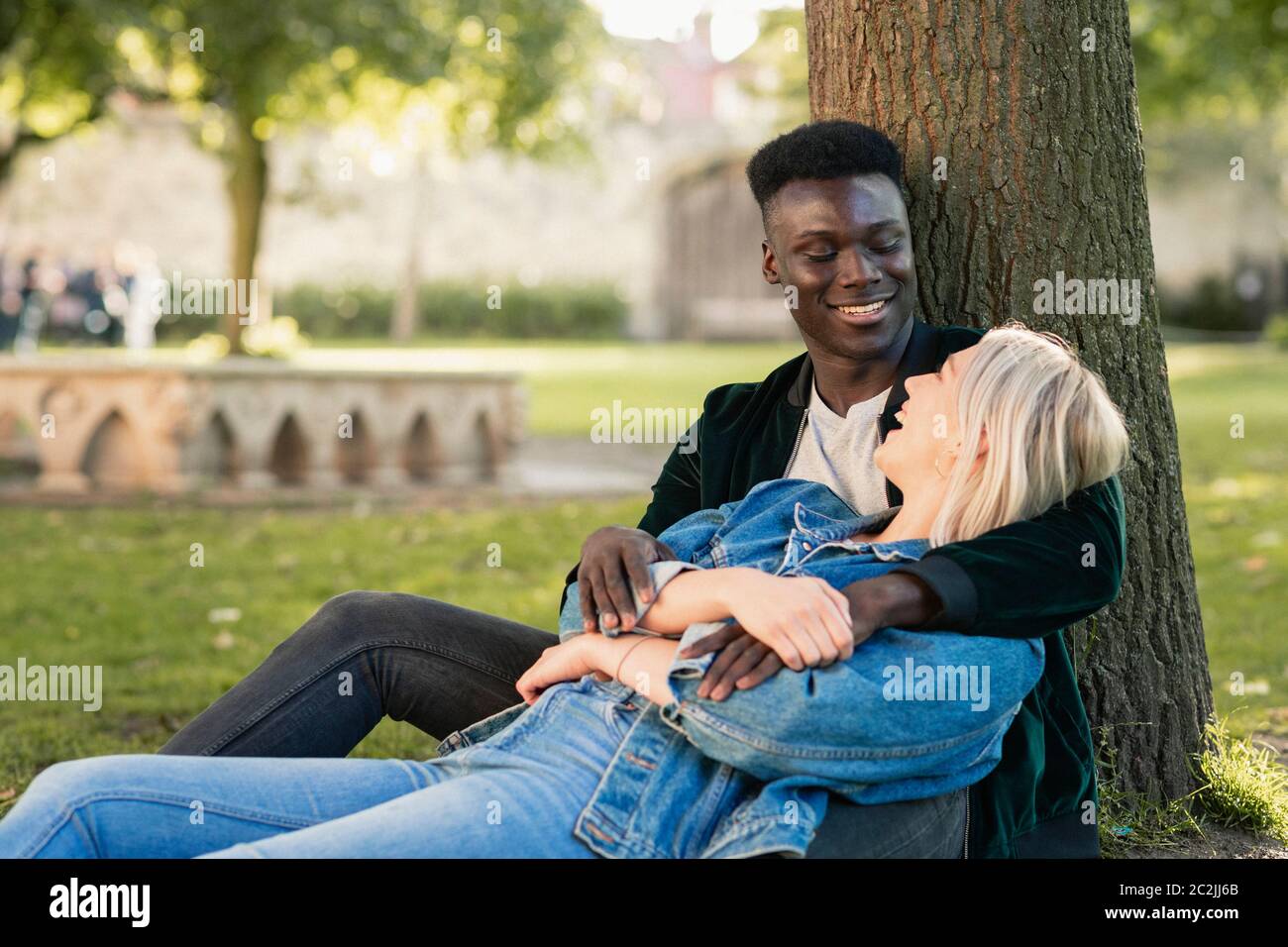 Ein Mann und eine Frau sitzen in einem Park gegen einen Baum. Der Mann ist, mit ihr, als sie zwischen seinen Beinen sitzt. Sie sind auf der Suche nach einander beim laughin Stockfoto