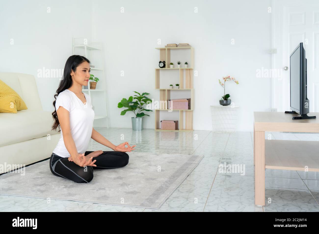 Schöne junge asiatische Frau in Sportbekleidung tun Yoga während im Wohnzimmer zu Hause entspannen, um Bewegung in ihren Alltag zu bekommen, während soziale di Stockfoto