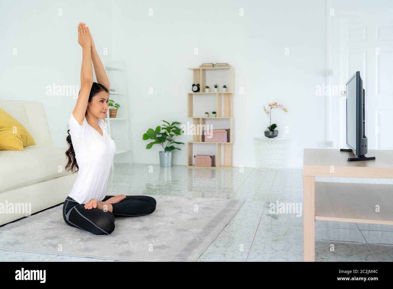 Schöne junge asiatische Frau in Sportbekleidung tun Yoga während im Wohnzimmer zu Hause entspannen, um Bewegung in ihren Alltag zu bekommen, während soziale di Stockfoto