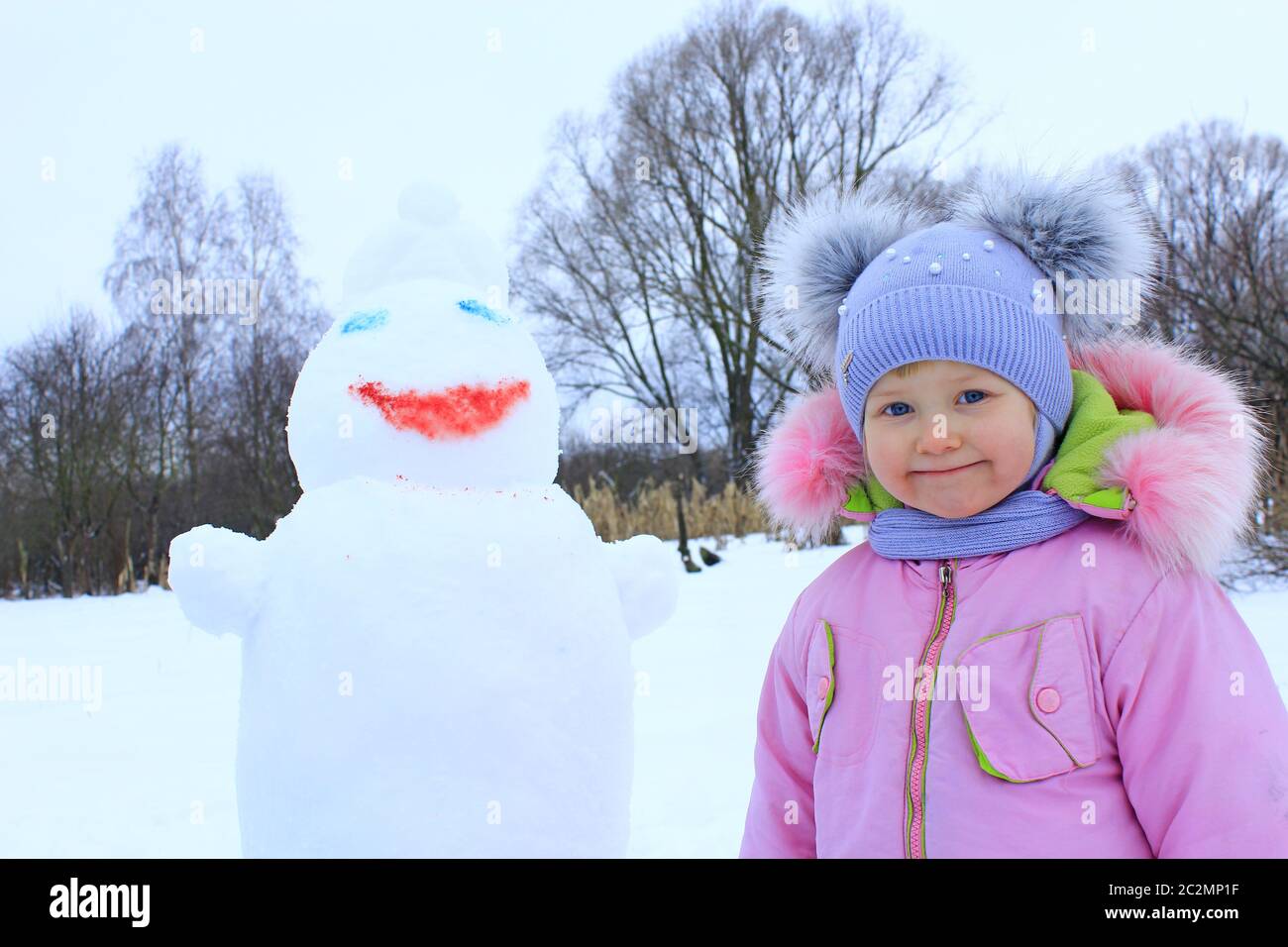 Porträt eines lächelnden Babys in amüsanten Wintermütze mit zwei lustigen Ohren. Kleines Mädchen und Schneemann Kopf Stockfoto