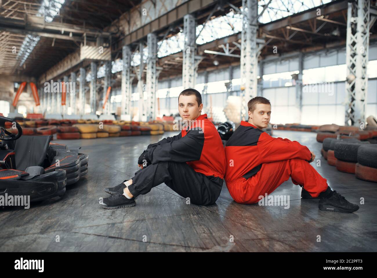 Zwei Kart-Rennfahrer sitzen Rücken an Rücken auf dem Boden, Kart Auto Sport Indoor. Speed Race auf engem Go-Kart Track mit Reifenbarriere. Schnelle Fahrzeugkompe Stockfoto