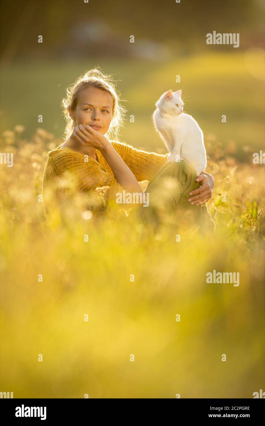 Hübsche, junge Frau mit ihrer Katze Pet im Gras sitzen auf der schönen Wiese beleuchtet durch warmes Abendlicht (flacher DOF; Farbe getonte Bild) Stockfoto
