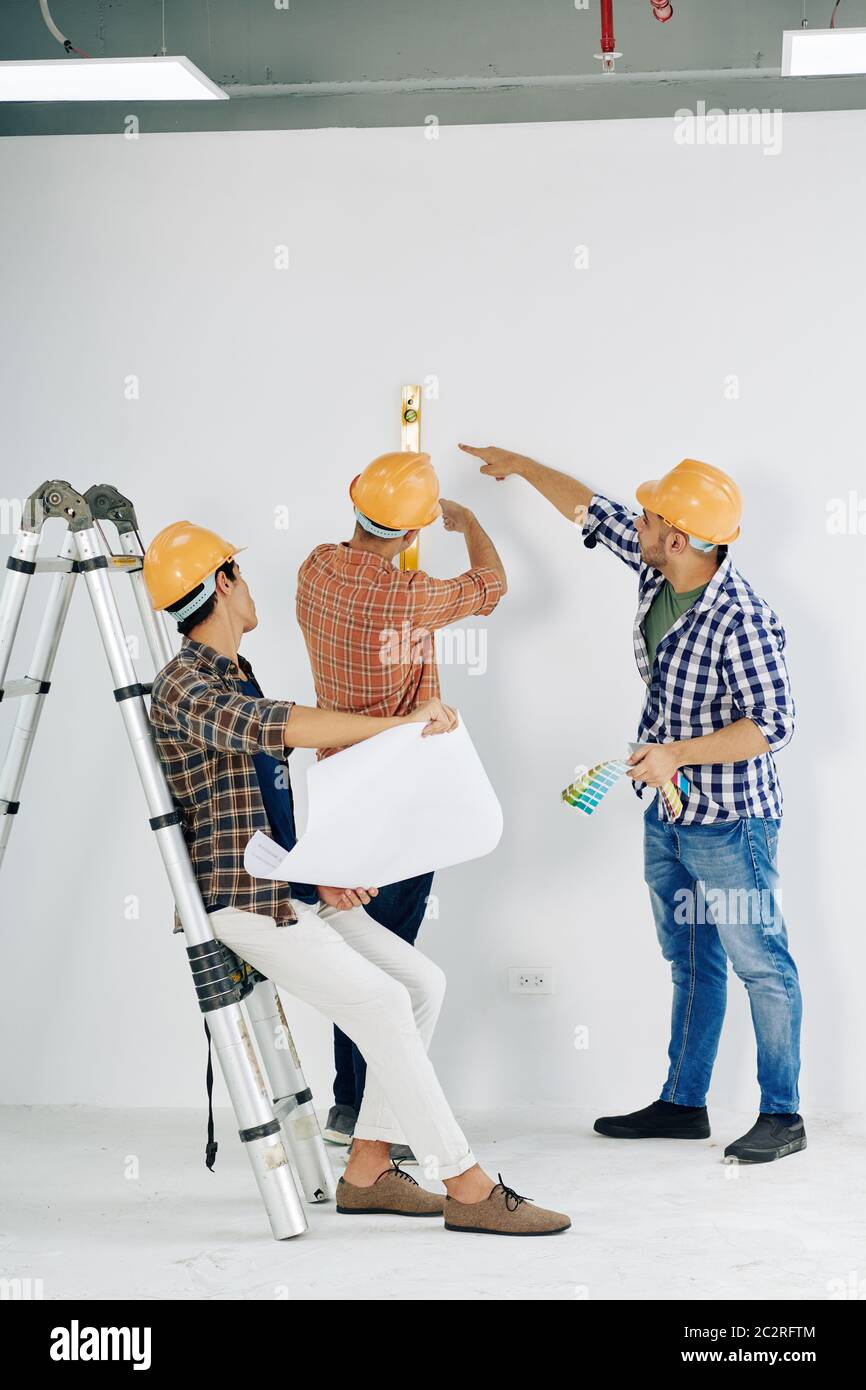 Moderne manuelle Arbeiter tragen gelbe Hardhats Überprüfung unfertigen Raum Wand Ebene, vertikale Aufnahme Stockfoto