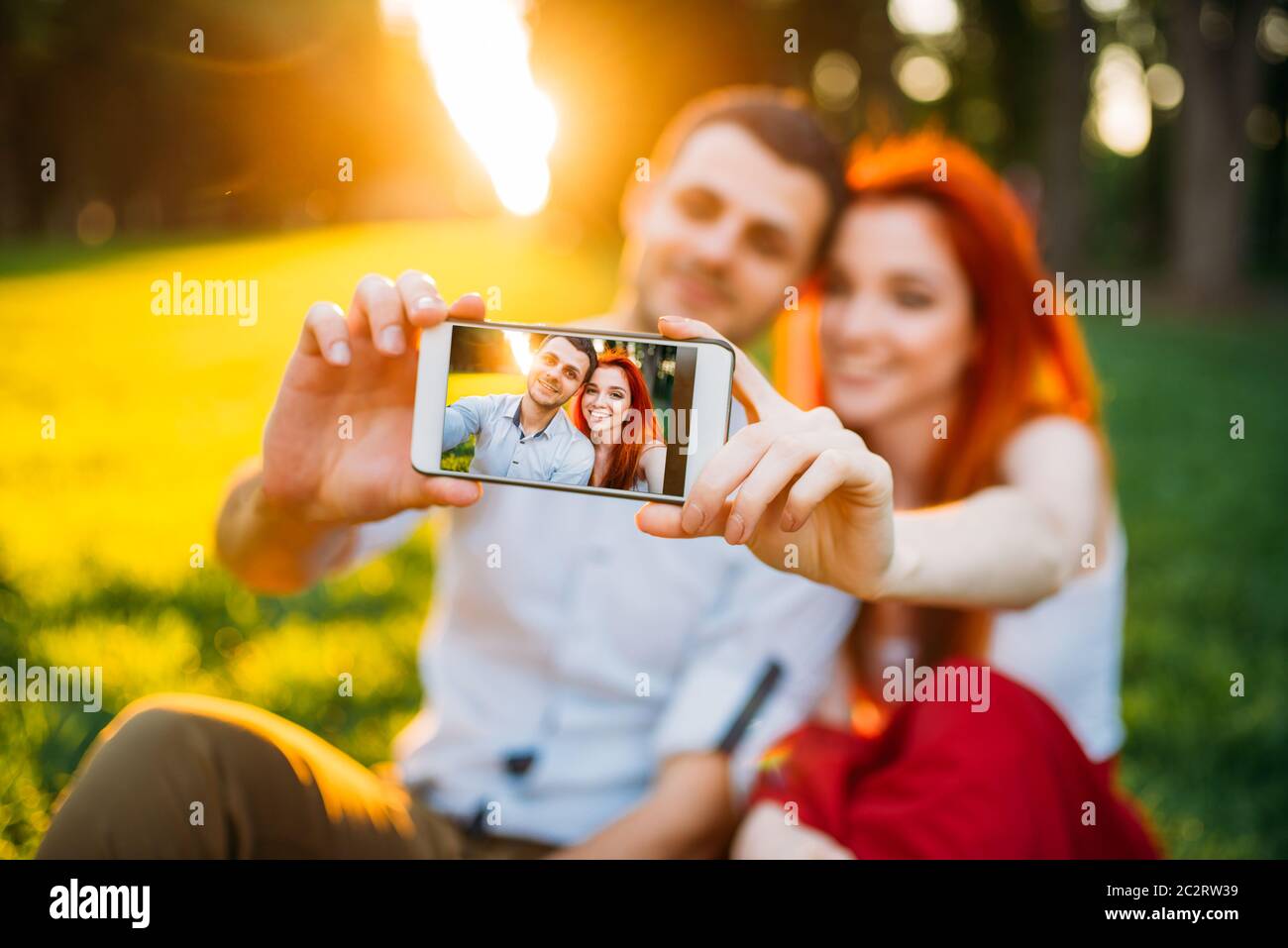 Liebespaar macht Selfie auf Handy-Kamera im Sommerpark am Sonnenuntergang. Romantisches Date attraktive Frau und junger Mann Stockfoto